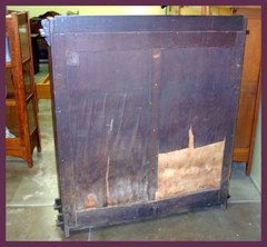 View of back, showing the paper label in the upper left hand corner and the moisture damage affecting only the floating panel of the frame and panel back. No damage to the panel's frame or the frame or feet of the bookcase.